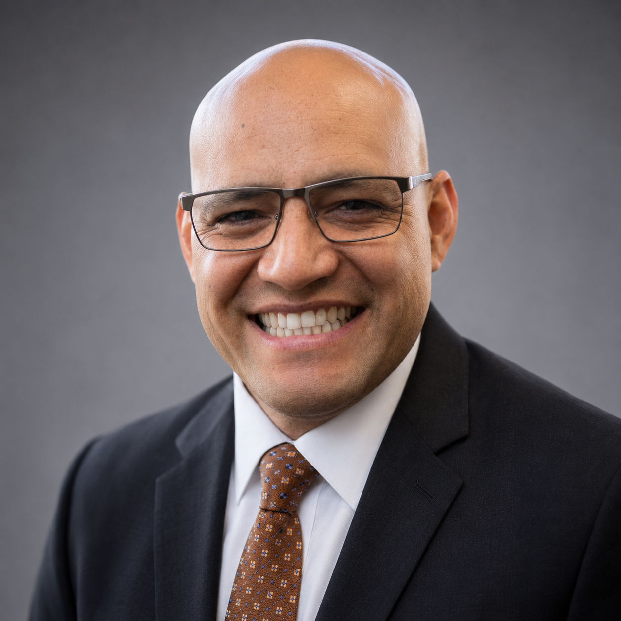 A man with a bald head and glasses is smiling, wearing a dark suit coat, white shirt and patterned tie.